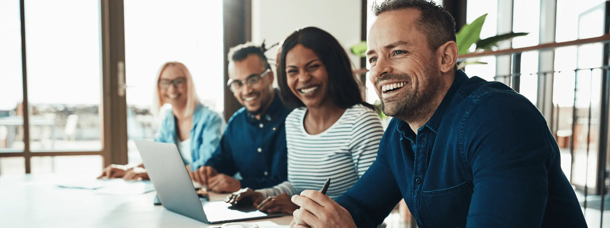 Four colleagues smiling around a meeting table with a laptop in a bright office
